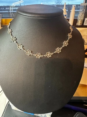 Silverfärgat blomhalsband - Ett tunt halsband i silverfärg med små, länkade blommor runt hela kedjan. Blommorna har en öppen design och ger ett lekfullt och stilrent intryck. Perfekt för dig som gillar detaljer