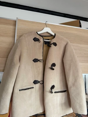 Beige fårskinnskappa från Zara - Snygg beige fårskinnskappa från Zara med dekorativa svarta knäppningar och detaljer i läderimitation. Rak modell med två fickor framtill och rundad halsringning. Perfekt för dig som vill ha en stilren och varm kappa med trendiga detaljer.
