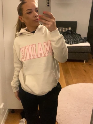 Vit hoodie med rosa MAINE-tryck - Säljer en vit hoodie med stor rosa MAINE-text på bröstet. Hoodien har huva och en klassisk magficka framtill. Perfekt för dig som gillar college-stilen och vill ha något mjukt och bekvämt. Materialet ser ut att vara bomullsblandning. Använd få tal gånger 