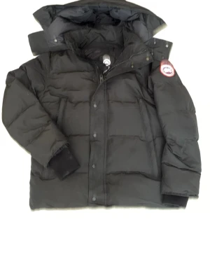 Svart dunjacka från Canada Goose - Säljer en svart dunjacka från Canada Goose med avtagbar huva och pälskant. Jackan har en klassisk patch på ärmen, dragkedja och knappar framtill samt ribbade muddar vid ärmsluten. Perfekt för kalla vinterdagar och riktigt varm.