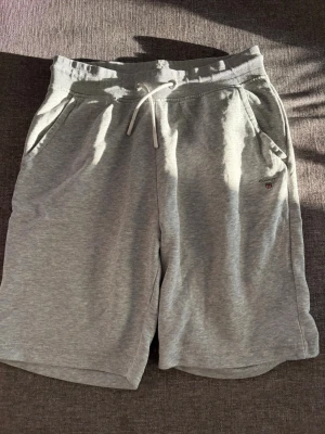 Grå mjukisshorts från Gant - Snygga grå mjukisshorts från Gant med dragsko i midjan och två sidofickor. Klassisk Gant-logga broderad vid fickan. Perfekta för chill dagar eller träning. Mjukt material som känns skönt mot huden.