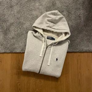 Grå zip hoodie från Polo Ralph Lauren - Snygg ljusgrå hoodie med dragkedja från Polo Ralph Lauren. Klassisk design med huva och vita dragsnören, samt den ikoniska lilla svarta logotypen broderad på bröstet. Tillverkad i mjukt bomullsmaterial för maximal komfort.