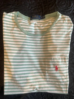 Randig t-shirt från Polo Ralph Lauren - Snygg randig t-shirt från Polo Ralph Lauren i ljusgrönt och vitt. Klassisk passform med rund halsringning och bröstficka med röd broderad logga. Perfekt för dig som gillar stilrena och fräscha plagg. Postas dagen eller dagen efter köpet.
