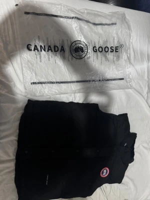 Svart dunväst från Canada Goose - Snygg svart dunväst från Canada Goose med klassisk logotyp på bröstet. Västen har tryckknappar framtill och är tillverkad i slitstarkt material med dunfyllning för extra värme. Perfekt för lager-på-lager och streetwear-stil.