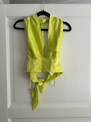 Limegrön omlottopp med knyt - Trendig limegrön omlottopp med bred midjedel och knyt både i nacken och ryggen. Toppen är ärmlös och har en djup v-ringning, perfekt för att sticka ut. Sval och lätt i syntetmaterial med snygg glans.