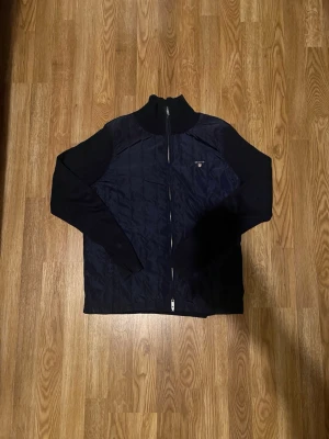 Marinblå quiltad cardigan från Gant - Snygg marinblå cardiganjacka från Gant med quiltat mönster och dragkedja framtill. Jackan har ribbstickad krage och ärmar samt Gant-logga broderad på bröstet. Perfekt för lager-på-lager och stilrena outfits. Även perfekt för lite kyligare sommarkvällar och för den som vill klä sig med stil och grisch!!