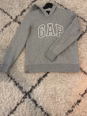 Grå hoodie från GAP med huva - Snygg grå hoodie från GAP med stor vit logga på bröstet. Hoodien har huva och en klassisk magficka framtill. 