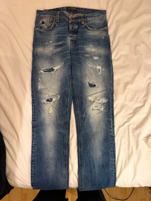 Blå slitna jeans från Nudie Jeans - Säljer ett par blå jeans från Nudie Jeans med tydliga slitningar och hål på framsidan för en riktigt avslappnad look. JEANSEN ÄR OMSYDDA TILL LÄNGDEN 34 och INTE 36