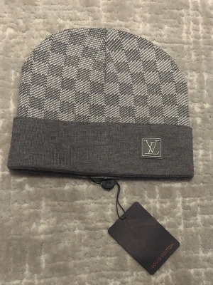 Grå Louis Vuitton mössa med mönster - Snygg grå mössa Louis Vuitton med klassiskt rutmönster och vikt kant. LV-logga framtill på en diskret patch. Tillverkad i mjukt stickat material som känns lyxigt och bekvämt. Perfekt accessoar för dig som gillar exklusiv streetstyle.