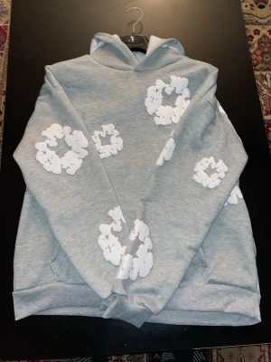 Grå denim tears hoodie - Säljer en grå hoodie med stora och små vita molnliknande tryck över hela tröjan. Hoodien har en klassisk huva och en stor magficka framtill. Tillverkad i mjukt sweatshirtmaterial som är perfekt för chill dagar. Fet fit för en avslappnad stil.