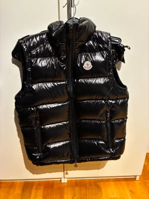 Svart dunväst från Moncler helt oanvänd - Snygg svart dunväst från Moncler med glansig finish, hög krage och huva. Västen har två dragkedjeförsedda fickor fram och Moncler-logga på bröstet. Perfekt för lager-på-lager och ger en cool streetwear vibe.