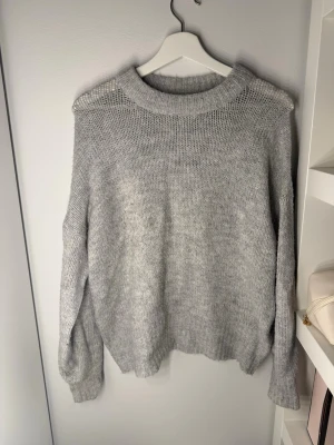 Ljusgrå oversized stickad tröja - Mysig ljus sopgrå stickad tröja med bred ribbad krage och avslappnad passform. Tröjan har långa ärmar och är tillverkad i ett mjukt, fluffigt material som känns skönt mot huden. Perfekt för lager på lager och chill dagar.