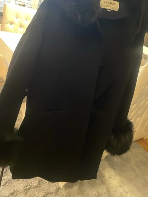 Svart kappa med fuskpäls från Hollies - Stilren svart kappa från Hollies med fuskpälsdetaljer vid krage och ärmslut. Kappan har raka fickor och en diskret guldfärgad logga på ärmen. Perfekt för dig som vill ha en klassisk look med en lyxig touch.