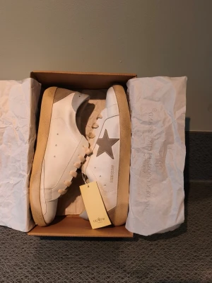 Golden Goose Superstar storlek 42 - Golden Goose Superstar sneakers i vitt läder med beige mockadetaljer och grå stjärna på sidan. Klassisk låg modell med beige sula och vita skosnören. Märkeslogga på plösen och hälpartiet. Snygg och trendig design för dig som gillar streetstyle. 