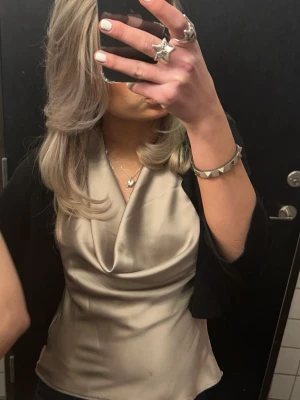 Champagnefärgad halterneck topp Zara - Säljer en champagnefärgad halterneck topp från Zara med draperad front och croppad passform. Toppens material är glansigt och känns som satin, perfekt för att skapa en snygg look. Passar dig som gillar stilrena och trendiga plagg.