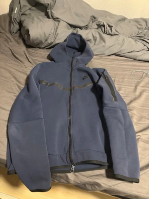 Blå hoodiejacka från Nike - Mörkblå hoodiejacka från Nike med dragkedja framtill och huva. Jackan har svarta detaljer längs bröstet och på ärmen, samt en ficka med dragkedja på ena ärmen. Perfekt för en sportig och avslappnad stil.