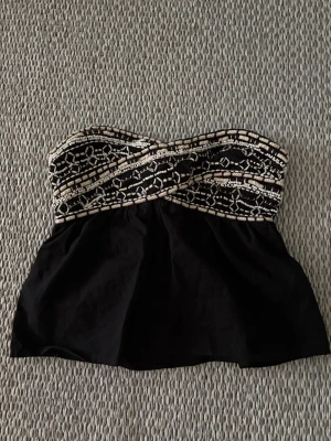 Svart axelbandslös topp  - Snygg svart bandeautopp med mönstrad överdel i vita och beigea mönster/pärlor. Toppen är axelbandslös och har en lätt utsvängd passform nedtill. Perfekt för dig som vill ha en cool topp till sommaren. 🌺🌤️Köpt på zara. Vissa pärlor/dekorationer har lossnat. 