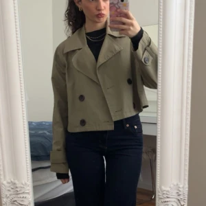 Beige croppad trenchcoat med knappar - Säljer en croppad trenchcoat med stora svarta knappar och bred krage. Jackan har långa ärmar med snygga detaljer vid ärmsluten och en loose passform. Perfekt för dig som vill ha en stilren och trendig jacka till våren eller hösten. Från Zara orginalpris 600 