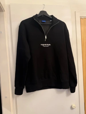Svart halvzip hoodie från Jack & Jones - Svart hoodie från Jack & Jones Originals med halvzip och broderad text 'Originals Studio' på bröstet. Tröjan har ribbade muddar och är gjord i mjukt material som känns skönt mot huden. Perfekt för dig som gillar en clean och stilren look. I mycket bra skick.