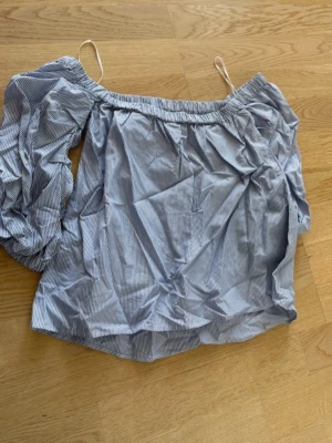 Blårandig offshoulder topp - Trendig blårandig offshoulder topp med puffig lång ärm på ena sidan och smala axelband. Croppad modell som ger en cool och avslappnad vibe. Perfekt för dig som vill sticka ut lite extra.