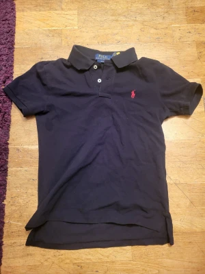 Svart pikétröja från Polo Ralph Lauren - Klassisk svart pikétröja från Polo Ralph Lauren med korta ärmar, krage och två knappar framtill. Tröjan har den ikoniska röda logotypen broderad på bröstet och är gjord i mjuk bomull för en bekväm känsla. Storlek 10-12 år. Snabb leverans