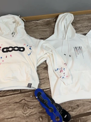 Vit hoodie med tryck från GOOD - Säljer en vit hoodie från GOOD med stor svart logga på bröstet och färgglada stänk i rosa och blått på ärmar och framsida. Hoodien har huva med dragsko och en stor magficka. Perfekt för dig som gillar streetwear och vill sticka ut lite extra.