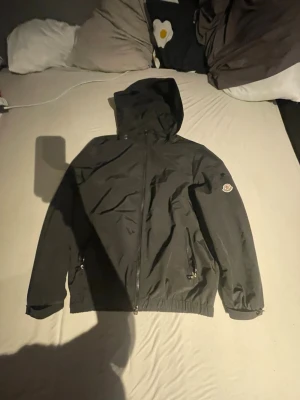 Svart vindjacka från Moncler - Svart vindjacka från Moncler med avtagbar huva och dragkedja framtill. Jackan har två sidofickor med dragkedja och Moncler-logga på vänster ärm. Materialet är lätt och vattenavvisande, perfekt för blåsiga dagar.