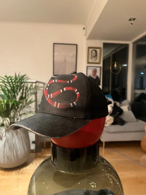 Svart keps med röd S-logga - Cool svart keps med stor S-logga i rött och vitt mönster på framsidan. Kepsen har böjd skärm och justerbar baksida. Perfekt för att ge din outfit en sportig touch.