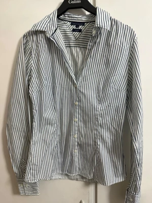 Randig skjorta från Tommy Hilfiger - Klassisk vit skjorta med smala marinblå ränder från Tommy Hilfiger. Skjortan har lång ärm, knappar framtill och en snygg krage. Tillverkad i bomull för en skön känsla och stilren look. Perfekt till jeans eller kostymbyxor.