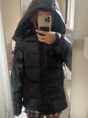 Svart pufferjacka - Säljer en svart pufferjacka med stor huva och dragkedja framtill. Jackan har en glansig finish, quiltad design och elastiska muddar vid ärmsluten. Perfekt för kalla vinterdagar och riktigt skön att bära. Passar dig som vill ha en stilren och varm jacka.