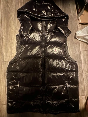 Svart dunväst från Moncler med huva - Snygg svart dunväst från Moncler med glansig finish och stor huva. Västen har dragkedja framtill, två fickor med dragkedja och Moncler-logga på bröstet. Perfekt för lager-på-lager och streetstyle.