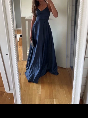 Marinblå långklänning med slits - Elegant marinblå långklänning med v-ringad omlott överdel och breda axelband. Klänningen har en markerad midja och en snygg slits på sidan som ger ett stilrent intryck. Perfekt för dig som vill ha en klassisk och tidlös look.