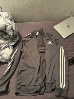 Grå Adidas träningsjacka - Säljer en grå träningskofta från Adidas med klassiska vita ränder längs ärmarna och logga på bröstet. Jackan har dragkedja framtill, ribbade muddar och är tillverkad i ett mjukt syntetmaterial. Perfekt för sport eller chill.