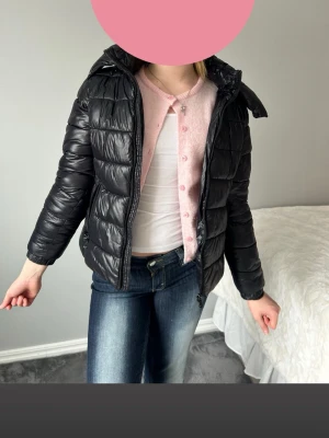 Svart pufferjacka med huva - Säljer en svart pufferjacka med huva och dragkedja framtill. Jackan har en klassisk quiltad design och är lätt vadderad för extra värme. Passar perfekt för kalla vinterdagar och har en stilren look som funkar till det mesta.