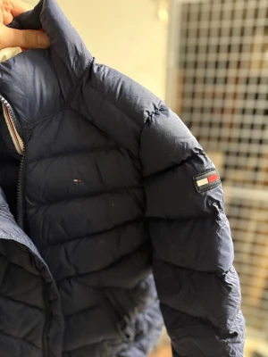 Mörkblå dunjacka från Tommy Hilfiger - Snygg mörkblå dunjacka från Tommy Hilfiger med klassisk quiltad design och dragkedja. Jackan har logotyp på ärmen och bröstet samt detaljer i rött, vitt och blått på insidan. Perfekt för kalla vinterdagar och riktigt stilren.