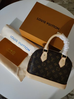 Iouis Vuitton handväska  - Butikspriset är 23 000kr.En elegant handväska i klassiskt Monogram-canvas med detaljer i ljust vachetta-läder. Väskan har den ikoniska strukturerade kupolformen och är utrustad med dubbla handtag samt en matchande clochette (nyckelhållare).