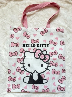 Hello Kitty shoppingväska rosa/vit - Supersöt shoppingväska i tyg med Hello Kitty-tryck, rosa handtag och massor av rosa rosetter och hjärtan. Väskan är vit med svart och rosa detaljer och har en klassisk fyrkantig form. Perfekt för dig som älskar kawaii och vill ha en unik accessoar till vardagen.