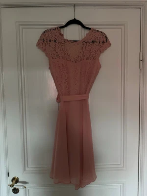 Rosa spetsklänning med bälte Esprit - Söt rosa kortklänning med spetsdetaljer och rund hals. Klänningen har ett matchande bälte i midjan och korta ärmar. Överdelen är helt i spets med blommigt mönster och kjolen är lätt utsvängd i ett tunt material.