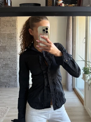 Svart skjorta med volangdetaljer - Säljer en svart skjorta med snygga volangdetaljer längs knäppningen. Alla knappar funkar orkade bara inte knäppa alla 😅 