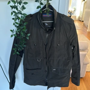 Olivgrön vindjacka från Snoot, storlek L - Olivgrön vindjacka från Snoot med coola detaljer som axelklaffar, fyra framfickor med knappar och dragkedja framtill. Jackan har en stilren look och är gjord i polyester, perfekt för dig som gillar funktion och snygg design. Passar dig som vill ha en jacka med lite edge.