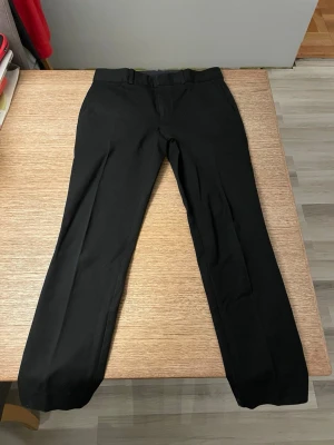 Svarta klassiska kostymbyxor från Ralph Lauren - Snygga svarta kostymbyxor med rak passform från Polo Ralph Lauren. Byxorna har diskreta fickor bak och är perfekta för en stilren look. Materialet känns mjukt och följsamt, vilket ger en bekväm känsla hela dagen.