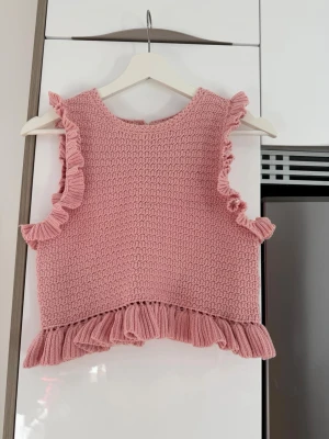 Rosa stickad croppad topp med volang - Söt rosa stickad topp med volangdetaljer vid ärmar och nederkant. Croppad modell som passar perfekt till jeans eller kjol. Toppen är ärmlös och har en mjuk, strukturerad stickning som ger en trendig look.