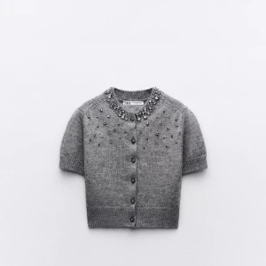 Grå kortärmad kofta med pärlor Zara - Kortärmad grå kofta från Zara med dekorativa pär tofsar och stenar runt halsringningen och axlarna. Koftan har rund hals, knäppning framtill och ribbade kanter. Prislapp kvar!! Nypris 260