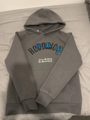 Hoodrich hoodie - Säljer en grå hoodie från Hoodrich med tryck i svart, blått och vitt på bröstet. Texten 'From Nothing To Something' står under loggan. Hoodien har huva, magficka och ribbade muddar. Perfekt för en avslappnad streetwear-look. Tröjan är väldigt fin och endast använd ett fåtal gånger. Kom dm om du är intresserad eller har frågor