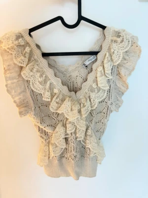 Beige stickad blus med spetsvolanger - Romantisk beige stickad blus med breda spetsvolanger längs v-ringningen och axlarna. Ärmlös modell med ett detaljerat mönster. Perfekt för dig som gillar en feminin stil🌷🪽Säljer även samma i svart!!!