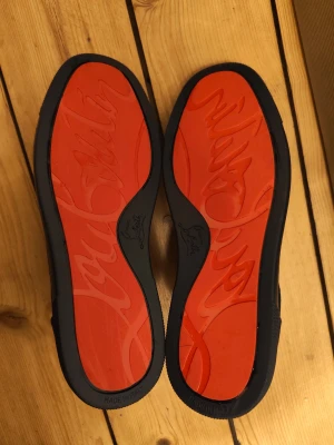 Mörkblå Christian Louboutin sneakers - Snygga mörkblå sneakers från Christian Louboutin med klassisk röd sula och diskret logga. Skorna har lågt skaft, svarta skosnören och ovandel i textil och läder. Perfekta för dig som vill sticka ut med exklusiv stil.