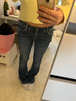 Mörkblå bootcut jeans med låg midja - Säljer ett par mörkblå bootcut jeans med låg midja och klassiska fem fickor. Jeansen har snygga slitningar och kontrastsömmar. Perfekta för dig som gillar en avslappnad och trendig stil.