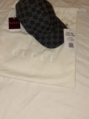 Gucci keps  - Snygg Kaschmir mörkgrå keps från Gucci i ullmix med det klassiska GG-monogrammet över hela kepsen. Justerbar baktill med kardborreband och svart insida. Kepsen kommer med original dustbag och taggar kvar. Perfekt för dig som vill ha en exklusiv och trendig accessoar.