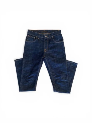 Mörkblå jeans från Nudie Jeans - ett par klassiska mörkblå jeans från Nudie Jeans med raka sopiga ben och femficksmodell. Jeansen har kontrastsömmar och en läderpatch med logga bak i midjan. Tillverkade i kraftig denim, perfekta för en avslappnad och stilren look. Jag har använt de ett fåtal gånger men använder dem inte längre. 