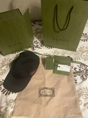 Svart keps från Gucci med röd-gröna detaljer - Snygg svart keps från Gucci med klassiska röd-gröna ränder på sidan. Kepsen har böjd skärm och justerbar rem baktill. Kommer med originalpåse, tags och Gucci-påsar. Perfekt accessoar för en trendig look. Kvitto, box allt ingår vid köp av kepsen.